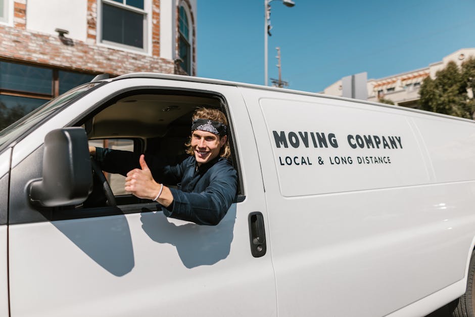 A young male mover with long hair, wearing a black bandana and a dark blue shirt, is sitting inside a white moving van with the driver’s side window open. He is smiling and giving a thumbs-up gestur