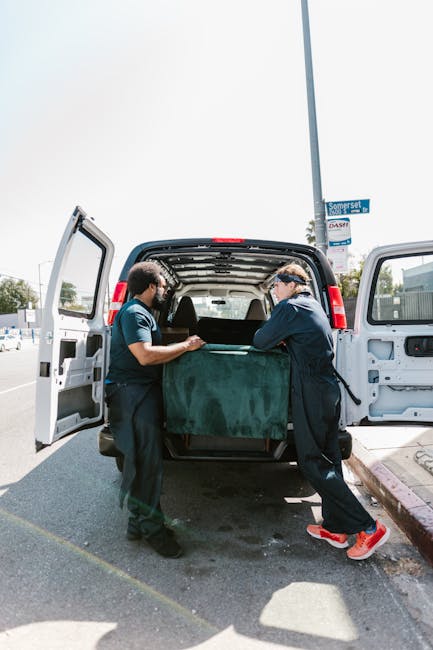 A young male mover with long hair, wearing a black bandana and a dark blue shirt, is sitting inside a white moving van with the driver’s side window open. He is smiling and giving a thumbs-up gestur
