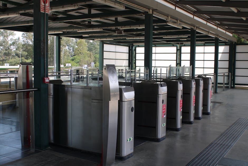 Inside a parking structure near Goodmayes Station, several stainless steel automated ticket barriers are aligned along a walkway, with a few glass panels and metal railings visible behind them. The barriers are equipped with card readers and sensors, some displaying red warning labels. Natural daylight filters through large, semi-transparent panels on the exterior walls, illuminating the concrete floor and the area around the barriers. Outside the open sides of the parking facility, trees and greenery are visible, indicating an outdoor environment beyond the covered area. This setting is relevant to home relocation and moving logistics, as it demonstrates a typical station parking scenario where vehicle access and passenger movement are facilitated for household removals, furniture transport, or professional moving services. The space appears clean and organized, with equipment such as trolleys, vans, or packing materials potentially used nearby, supporting efficient loading and transportation during the moving process performed by companies like manwithvangoodmayes.co.uk.