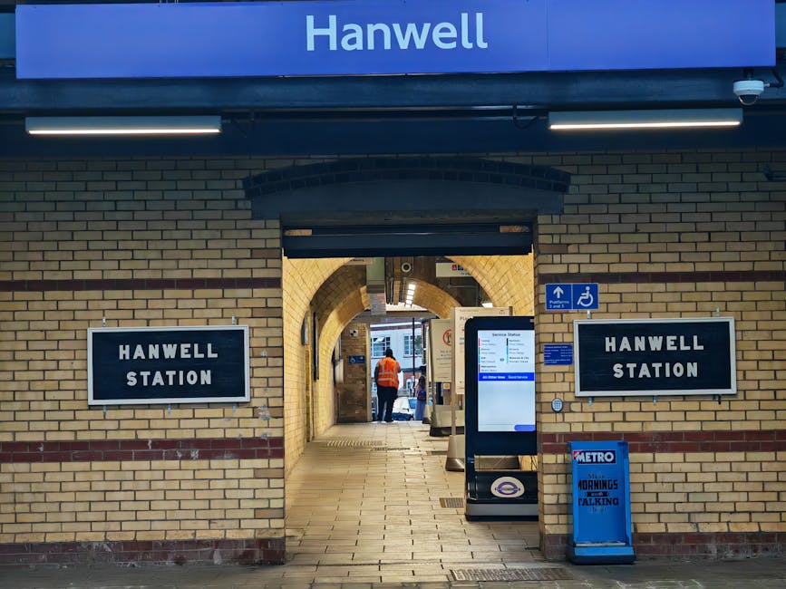 Interior view of Hanwell Station showing a brick archway leading to the platform entrance, with two black and white signs displaying 'HANWELL STATION' on either side of the passage. In the foreground, there is a digital information kiosk and a blue Metro card sales and travel information display. To the right, a wheelchair accessible sign is mounted on the brick wall. Inside the passage, a staff member wearing an orange high-visibility vest is visible, possibly assisting with home relocation or moving services associated with the station. The scene is staffed with station lighting and the environment appears clean and organized, supporting efficient passenger and furniture transport during packing and moving activities.