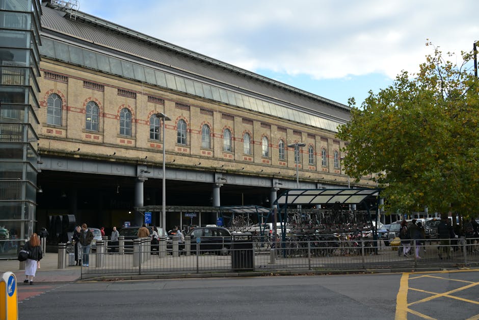 Inside a parking structure near Goodmayes Station, several stainless steel automated ticket barriers are aligned along a walkway, with a few glass panels and metal railings visible behind them. The barriers are equipped with card readers and sensors, some displaying red warning labels. Natural daylight filters through large, semi-transparent panels on the exterior walls, illuminating the concrete floor and the area around the barriers. Outside the open sides of the parking facility, trees and greenery are visible, indicating an outdoor environment beyond the covered area. This setting is relevant to home relocation and moving logistics, as it demonstrates a typical station parking scenario where vehicle access and passenger movement are facilitated for household removals, furniture transport, or professional moving services. The space appears clean and organized, with equipment such as trolleys, vans, or packing materials potentially used nearby, supporting efficient loading and transportation during the moving process performed by companies like manwithvangoodmayes.co.uk.