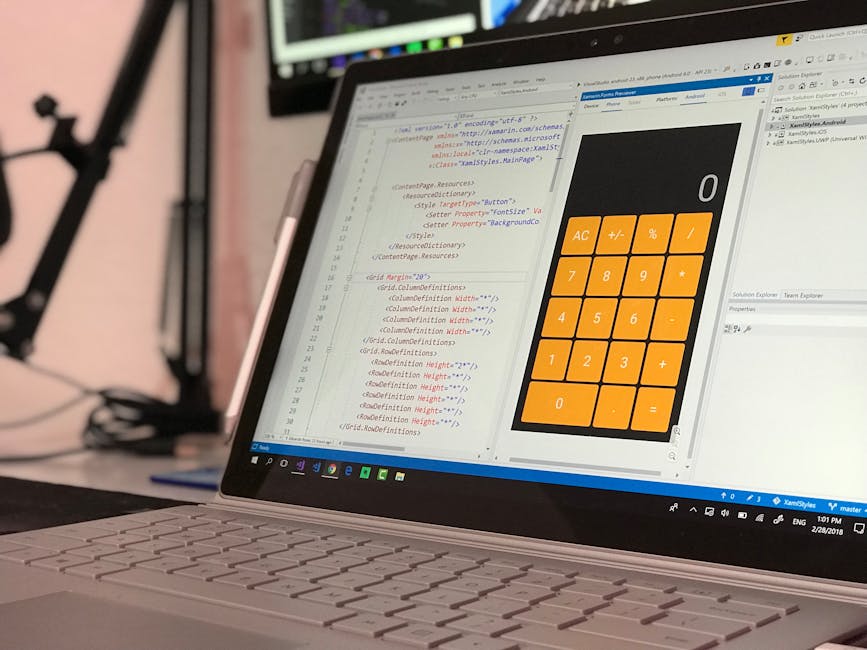 A close-up view of a laptop on a desk, displaying a programming environment with lines of code written in HTML and CSS on the left side of the screen, and a large, orange calculator application with white symbols on the right side. The background includes a blurred black tripod and other equipment, indicating a workspace setup, possibly for developing or managing home relocation and moving services. The scene reflects a professional and organized environment suitable for planning moves, packing, or calculating quotes related to removals, with natural lighting illuminating the workspace.