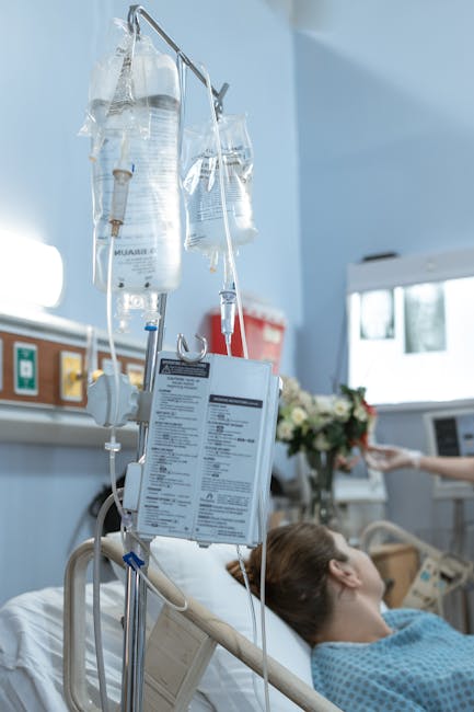 A patient's hospital bed in a clinical setting, with an intravenous (IV) drip stand positioned beside it. The IV drip bag, filled with clear fluid, is hanging from the metal pole with attached tubing leading to the patient. The patient, a woman with brown hair tied back, is lying on her side or back, wearing light blue hospital attire, and appears to be resting or waiting. In the background, medical equipment, a window allowing natural light, and a tray with flowers can be seen, indicating a well-maintained hospital room. The IV setup and bed are part of a typical hospital environment supporting patient care and treatment procedures related to hospital transfers or relocations.
