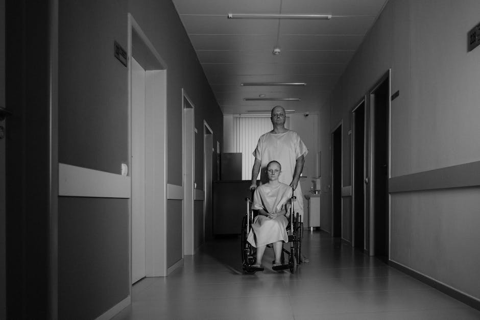 A black and white photograph depicts a hospital corridor with smooth, reflective flooring and ceiling mounted lights. Two individuals, both in patient gowns, are positioned centrally within the frame. One is seated in a wheelchair, with hands resting on the armrests, and appears calm. The other, standing behind, is pushing the wheelchair. The corridor is lined with several closed doors on either side, each with small signs above them. The far end of the corridor features a window with vertical blinds, allowing light to enter. Visible in the scene are hospital equipment, including a trolley and some boxes or packaging materials, suggesting a moving or relocation process within the hospital setting. The overall scene captures the typical environment of a healthcare facility, emphasizing the logistical aspects of patient transport or hospital moves, as could be relevant to home or institutional relocation services provided by manwithvangoodmayes.co.uk.