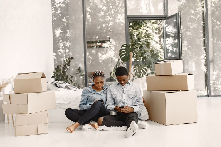 A young woman and a young man sit on the floor inside a bright, modern living room surrounded by packed cardboard moving boxes and plastic-wrapped furniture, indicating a home relocation or moving process. The woman has braided hair and is wearing a light blue denim jacket with black jeans, while the man, with short dark hair, is dressed in a light grey denim shirt and black trousers. Both of them are engrossed in their smartphones. Behind them, a large window and glass door allow natural light to illuminate the space, revealing indoor plants and a cozy, minimalist environment. The scene depicts the packing and moving stage before transportation, with the boxes and furniture prepared for loading onto a removal van, consistent with professional house removal services by companies such as manwithvangoodmayes.co.uk, for tenants and landlords managing tenancy move-outs.