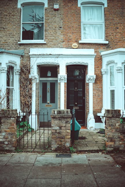 Photograph of two adjacent terraced houses on a residential street, with the ground-floor entrances visible. The house on the left has a blue door with a narrow vertical window, a small front garden enclosed by a low brick wall and a black metal gate, and a lantern-style light fixture hanging above the door. The house on the right features a black door with a gold handle and a door knocker, also with a small front garden enclosed by a low brick wall and a black metal gate. Both houses have large sash windows on the upper floors with white frames, and some windows have lightly drawn curtains. The brickwork of both houses is reddish-brown, with peeling white paint on the window sills and door frames indicating weathering. A narrow paved pathway leads to each front door, with some green plants and dried vines visible around the base of the left house. The overall scene depicts typical residential housing, likely within a London suburb, and is relevant for context regarding house removals and property moving services offered by companies such as manwithvangoodmayes.co.uk.