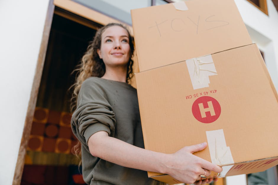 A young woman with curly hair, wearing a grey sweatshirt, is inside a house near a doorway and is carrying two cardboard boxes stacked vertically. The top box is labeled 'TOYS' written in pencil, while the larger box beneath has red printed markings and a red 'H' symbol, indicating fragile or hazardous contents. The boxes are taped securely with clear packing tape. In the background, a wooden door frame and some wall-mounted decor are visible, and the scene is lit with natural indoor lighting. This image illustrates the process of packing and moving household items, aligning with house removals and furniture transport services offered by manwithvangoodmayes.co.uk, as part of a home relocation or estate-specific move in the IG3 area.