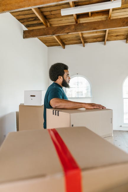 A man with dark curly hair and a beard, wearing a blue t-shirt, is standing inside a spacious room with white walls and a wooden ceiling with exposed beams. He is resting his hands on top of a large cardboard moving box, which appears to be sealed with packing tape. In the foreground, several other cardboard boxes are visible, some with red straps for securing items during a house move. Natural light enters through two arched windows on the right side of the image, illuminating the space. The room has a clean and minimalistic appearance, suitable for home relocation preparations. This scene depicts a professional packing and moving process, likely carried out by [COMPANY_NAME], focusing on efficient furniture transport and logistics for a house move in Goodmayes, IG3, as part of estate-specific removal services offered by [manwithvangoodmayes.co.uk].