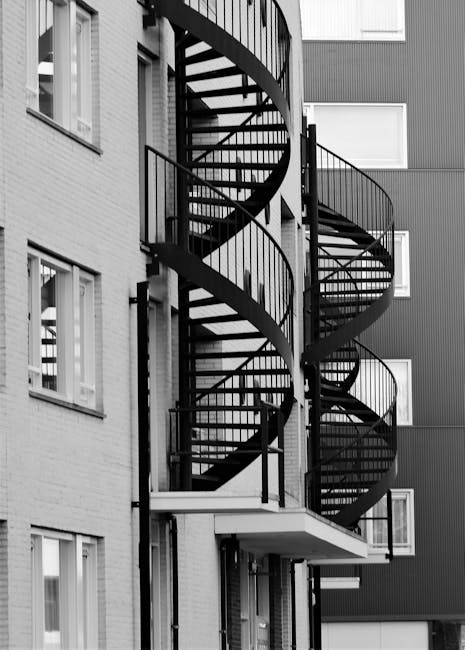 A black metal spiral staircase attached to the exterior of a multi-storey residential building in Goodmayes, featuring open risers and safety railings curving around each level. The staircase is positioned against the building's brick and panel facades, with multiple landings visible. The ground floor area includes a small canopy over the entrance and a tiled floor, with the staircase ascending to the upper floors, facilitating access for residents. This scene captures the typical layout for multi-storey housing in the area, illustrating a straightforward exterior access route that may require careful navigation during a home relocation or moving process. The lighting is natural, highlighting the structural details of the staircase and building features, reflecting a professional approach to property access for furniture transport and packing during removals, as handled by companies like manwithvangoodmayes.co.uk.