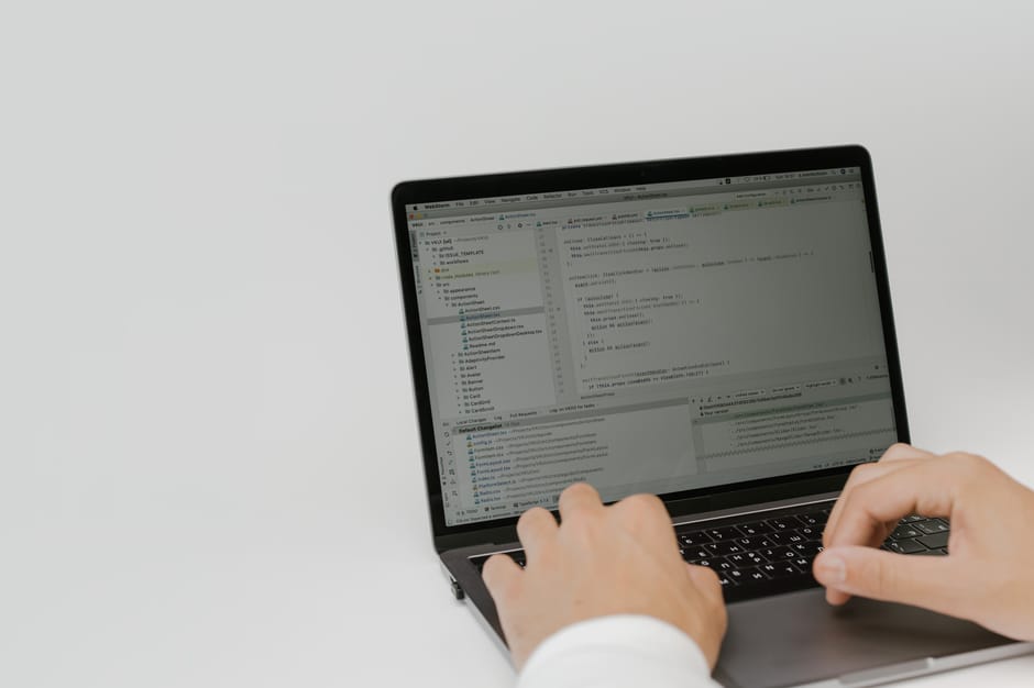 A person working on a laptop with a dark-colored keyboard and slim profile, displaying a code editor with multiple open tabs and lines of programming code on a bright, plain background. The individual's hands are positioned on the keyboard, and they are wearing a white long-sleeved shirt. The scene suggests a focus on digital or technical work, with no physical objects related to house removals visible in the image. This image represents a professional or administrative aspect of a home relocation or moving service, such as planning or documentation, aligned with the context of house removals and logistics by manwithvangoodmayes.co.uk.