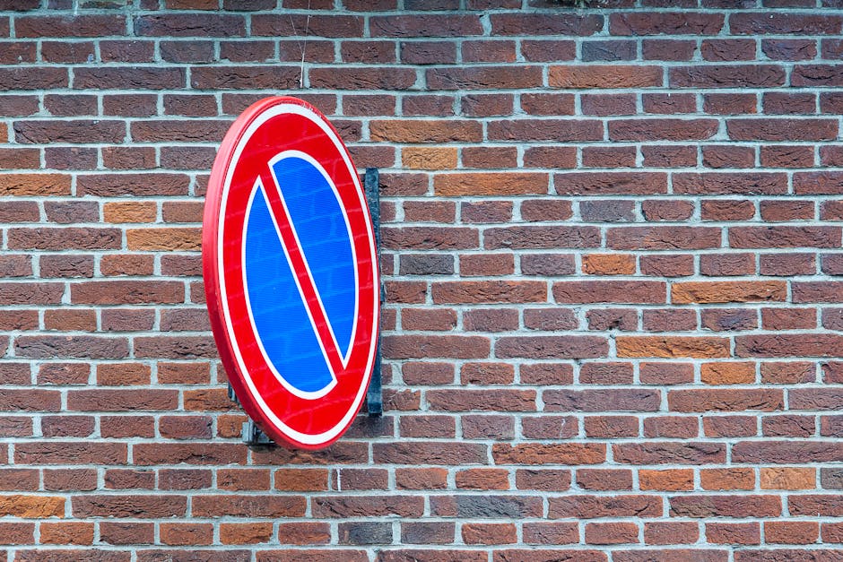 A round no parking traffic sign with a red border and a blue background featuring a red diagonal line crosses over a parking symbol, mounted on a black metal bracket attached to a red brick wall. The brick wall comprises evenly laid bricks with variations in shades of red, brown, and orange, and light mortar joints. The sign is positioned slightly to the left of the center in the image, with a clear, well-lit environment indicating daytime. This setting is part of a property where house removals or home relocation services may involve parking restrictions, relevant to logistics and transportation planning for removals. The image emphasizes traffic control measures that may impact loading and unloading of furniture and boxes during the moving process, reflecting parking and traffic regulations that [COMPANY_NAME] must consider for efficient removals in IG3 areas.