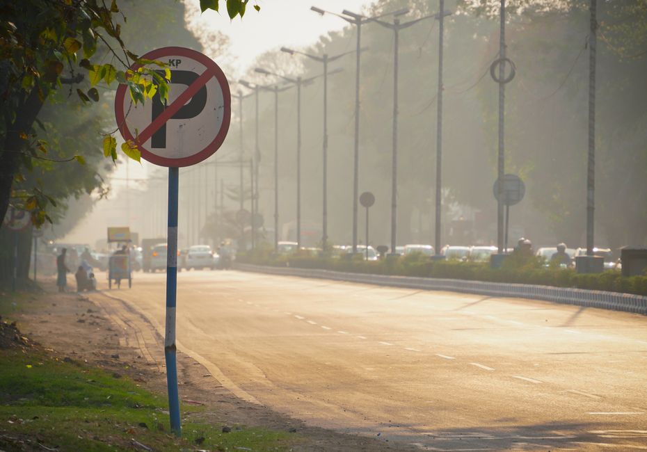 A no parking traffic sign with a red circle and diagonal line over a black letter 'P' is mounted on a blue and white striped pole along a paved asphalt street. In the background, there are several vehicles, including cars and motorcycles, moving along the road which is bordered by a curb and a sidewalk. The sidewalk is lined with trees whose green leaves partially shade the area, and a few pedestrians can be seen walking in the distance. Overhead, utility poles and power lines run parallel to the street, and the scene is illuminated by soft natural light, possibly early morning or late afternoon. This urban environment reflects typical parking regulations in a residential or commercial area, which is relevant for house removals and moving services planning in the IG3 postcode district. The image provides a clear view of the street environment where vehicle parking restrictions are in effect, supporting the logistical considerations for efficient home relocation or furniture transport by companies like manwithvangoodmayes.co.uk.