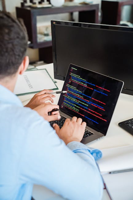 A person in a light blue shirt is seated at a desk in a well-lit room, working on a laptop displaying lines of colorful code on the screen. Behind the individual, there is a larger desktop monitor turned off, a white book or folder, and a black remote control. The desk appears to be located inside a home or office environment, with shelving and other furniture visible in the background. This scene illustrates a workspace related to packing, planning, or managing the logistics of household packing and moving services, aligned with the context of house removals and property relocation proposed by manwithvangoodmayes.co.uk. The person is using a pen to point at the laptop screen, indicating active engagement with the work involved in the moving process.
