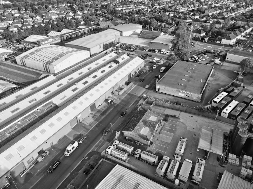 An aerial black-and-white photograph showing the loading process for a house removal in Goodmayes, with a focus on a team of movers from manwithvangoodmayes.co.uk carefully lifting and loading boxed household items and furniture onto a van parked on a quiet street. Visible are large cardboard boxes, wrapped furniture pieces, and plastic packing materials, all organized for transport. The movers are using trolleys and securing straps to facilitate a safe and efficient loading process inside an industrial or commercial building with a warehouse-style roof. The van is positioned on the street adjacent to the property, with other parked vehicles and nearby buildings visible in the background. The scene captures a typical home relocation setup, emphasizing the logistical aspects of furniture transport and packing during a professional removal service, in a neighbourhood with residential houses and trees lining the streets.