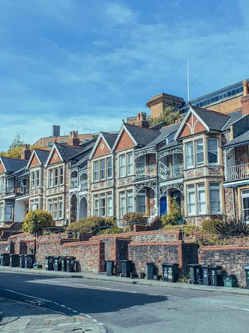 Image depicting a row of Victorian-style terraced houses with decorative white ironwork on their balconies and large bay windows, situated above a brick retaining wall along a residential street. The foreground shows a paved road with several black wheelie bins lined up along the curb and a sidewalk, while the background features a blue sky with some scattered clouds and trees behind the houses. This setting illustrates a typical UK home environment suitable for house removals and furniture transport services by manwithvangoodmayes.co.uk, with a focus on residential building exteriors and urban street scene.