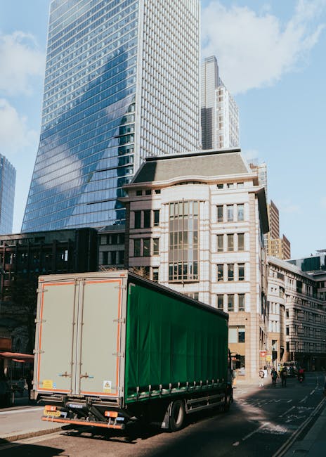 A large green delivery truck with a closed cargo area is parked on a city street in front of tall modern buildings featuring glass facades and older architectural structures. The truck is positioned near the curb, with its rear doors securely closed, likely in the process of loading or unloading household or office furniture during a home or office relocation. The street is busy with pedestrians and other vehicles, and the scene is illuminated by daylight, with shadows cast by the surrounding skyscrapers. This image illustrates urban logistics involved in furniture transport and packing for house removals, typical of professional moving services such as those offered by manwithvangoodmayes.co.uk, supporting efficient local or city-wide relocation or delivery operations.