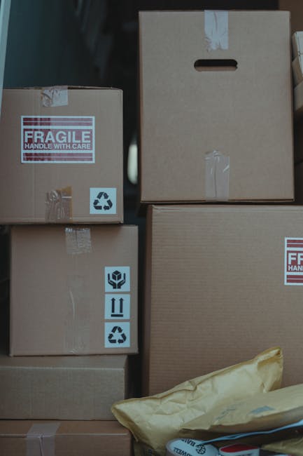A close-up view of several stacked cardboard moving boxes inside a property, prepared for a home relocation. The boxes vary in size and are sealed with clear packing tape, some featuring handling labels such as 'fragile' and recycling symbols. One box has a cut-out handle, indicating it is designed for easier lifting. A yellow padded envelope and a few loose items are visible at the base of the stack, suggesting packing and moving preparations. The environment appears to be an indoor storage or packing area, with the boxes arranged in an organized manner for loading onto a vehicle. The lighting is neutral, highlighting the textures of the cardboard and packing materials. This scene reflects the logistical aspects of furniture transport and packing involved in professional removals services, such as those offered by manwithvangoodmayes.co.uk under their house removals and urgent same-day moving solutions.